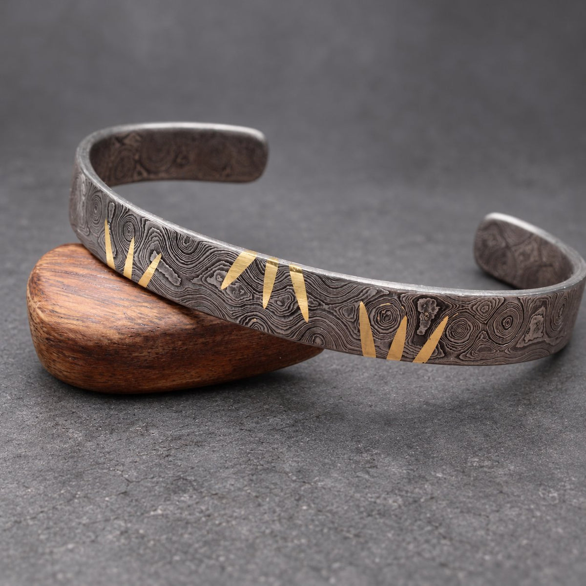 A dark gray metal cuff bracelet with a textured pattern and gold-colored accents. The bracelet is open-ended and rests on a wooden surface. The gold accents are in a repeating triangular design.
