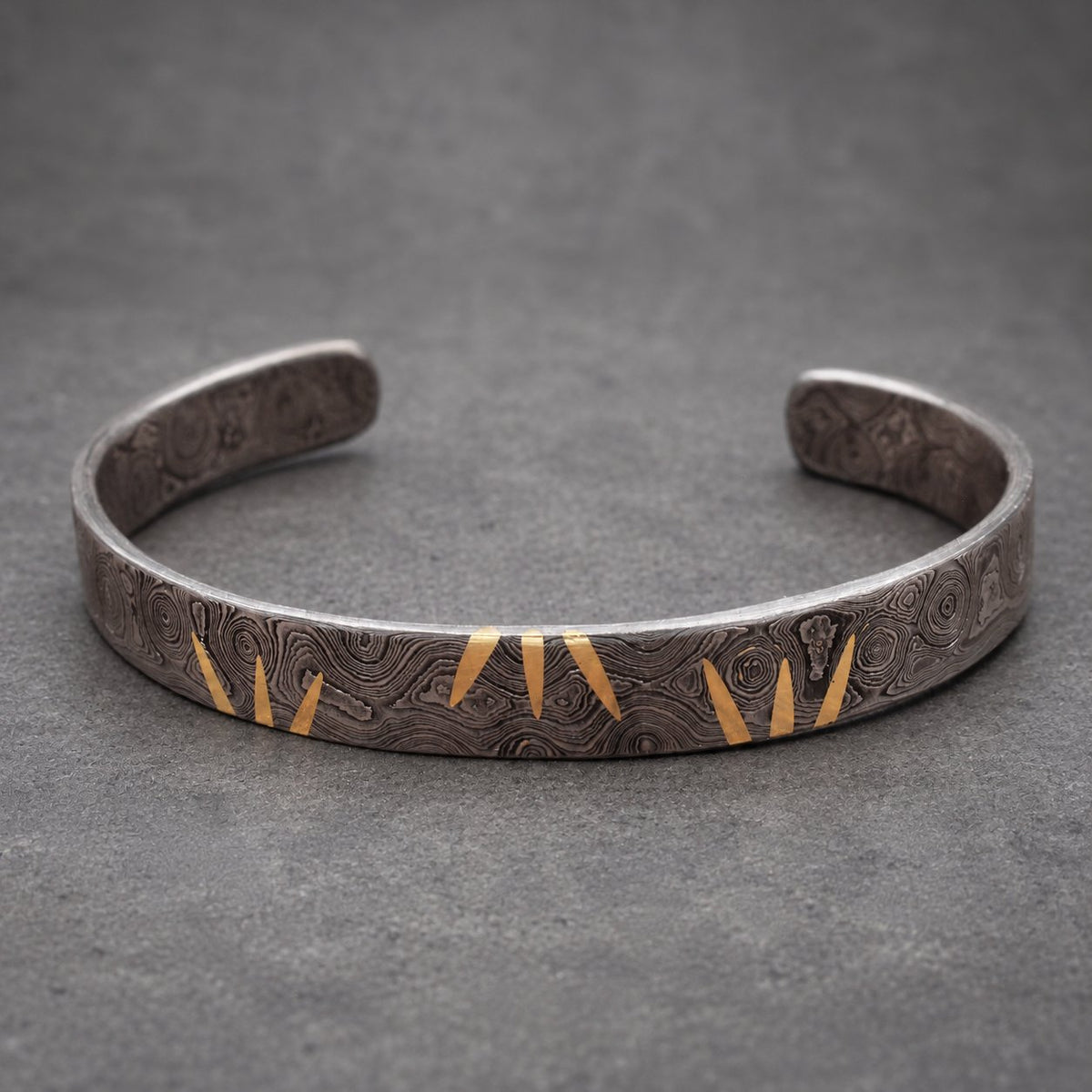 A silver-toned cuff bracelet with a textured pattern and gold-colored chevron accents. The open-ended bracelet is displayed on a wooden surface, showcasing its design. This piece is a stylish accessory.