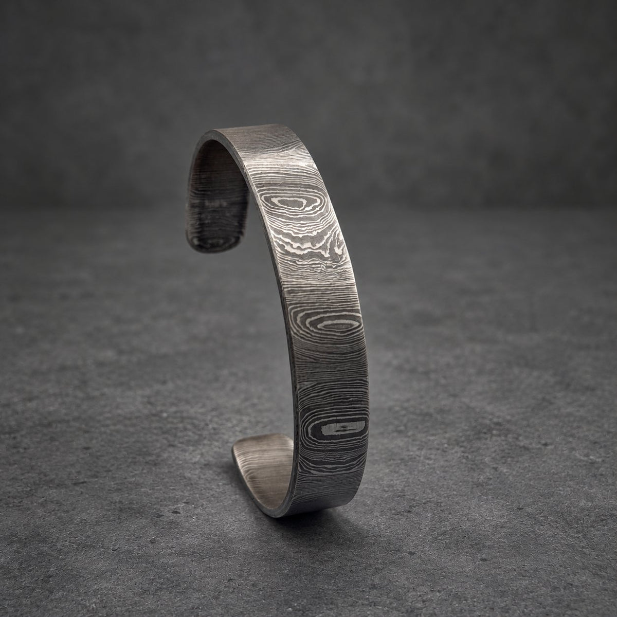 A Damascus steel cuff bracelet with a distinctive wood grain pattern. The open cuff design features a dark gray and silver color scheme. The bracelet is displayed on a gold-colored decorative stand against a gray background.