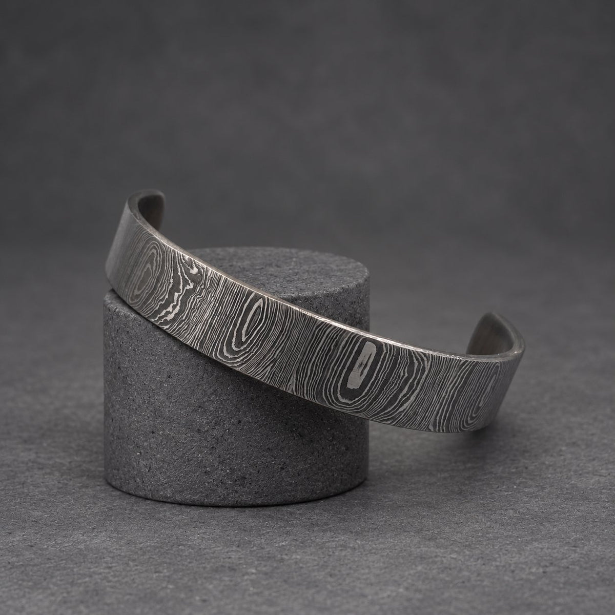 A dark gray metal cuff bracelet with a distinctive wood grain pattern. The bracelet is displayed on a gold-colored decorative stand. The cuff has a simple, open design.