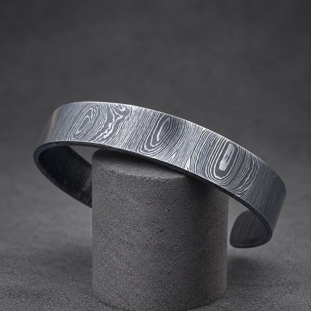 A silver-toned metal cuff bracelet with a distinctive wood grain pattern. The bracelet is open at the bottom and is displayed on a gold-colored, textured branch. The bracelet's surface has a swirling, layered design.