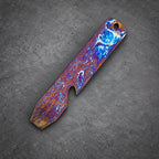 A rectangular bottle opener with a hole at one end. The opener has a striking pattern of blue, orange, and purple colors. The metal tool features a bottle-cap opener on one side.