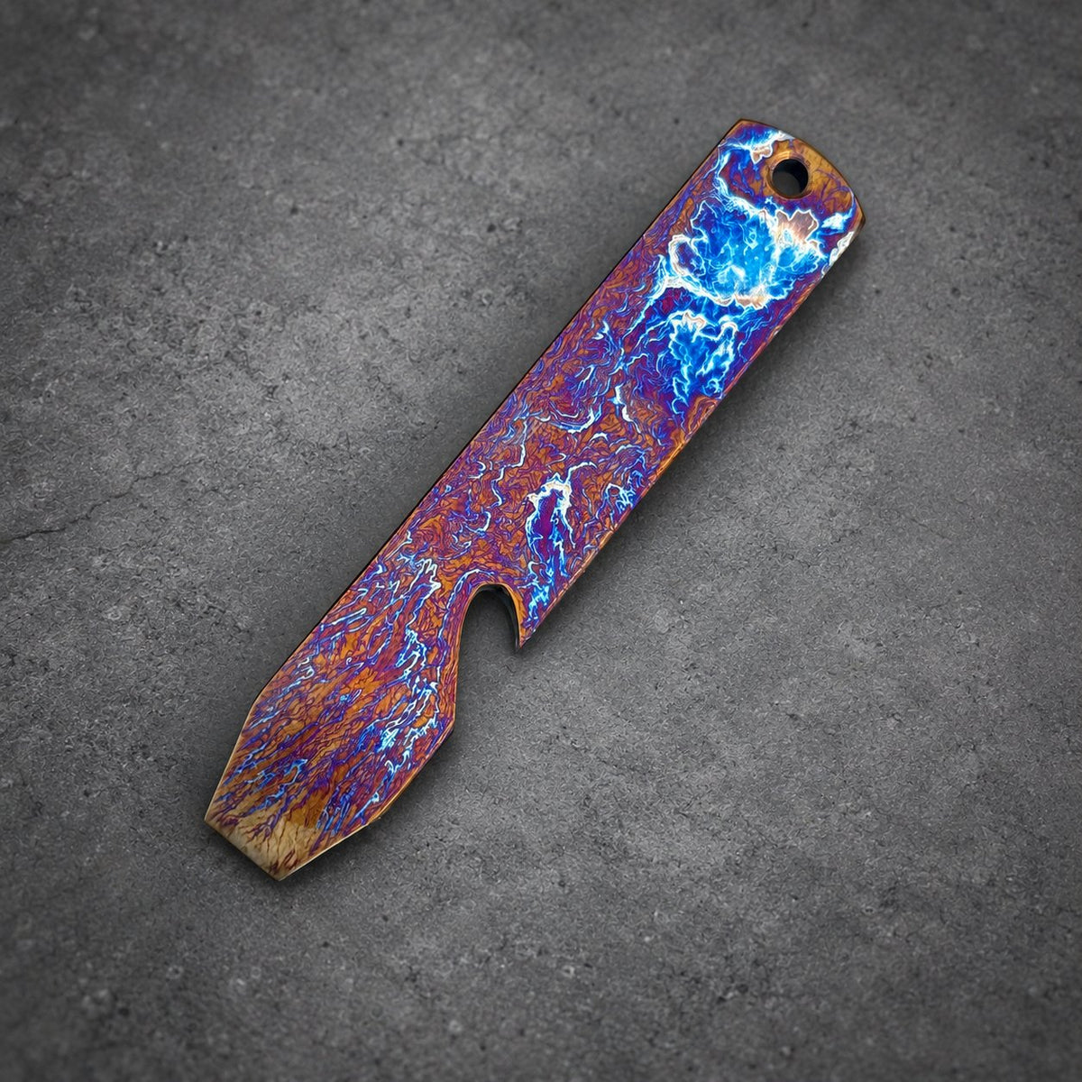 A rectangular bottle opener with a hole at one end. The opener has a striking pattern of blue, orange, and purple colors. The metal tool features a bottle-cap opener on one side.