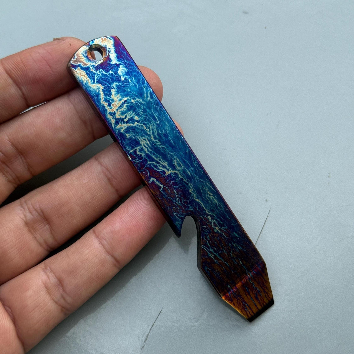 A multi-colored, rectangular bottle opener with a key ring hole. The opener features a blue, purple, and gold marbled design. The bottom of the opener has a bottle-opening notch.