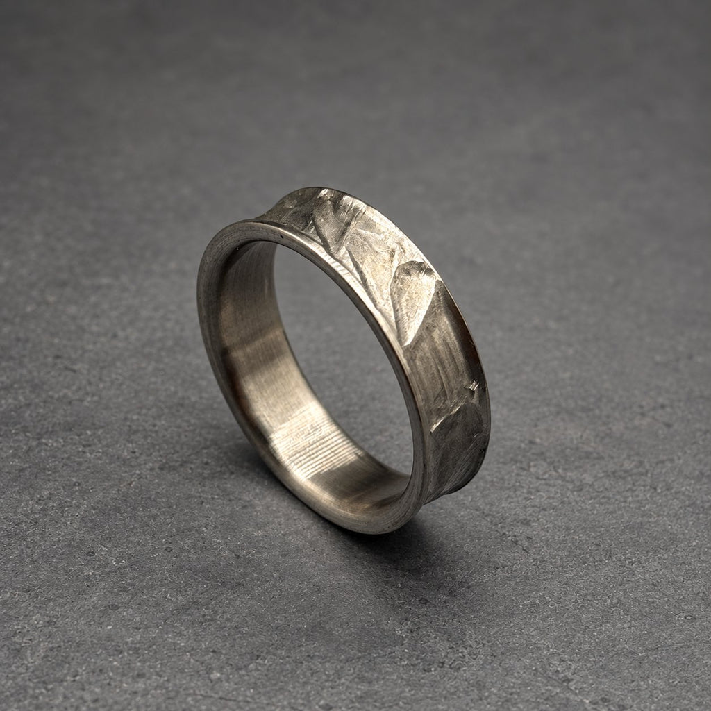 A silver ring with a textured surface. The ring has a slightly curved design and a band with a hammered or carved pattern. The ring is displayed on a gray surface.