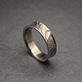 A silver ring with a textured surface. The ring has a slightly curved design and a band with a hammered or carved pattern. The ring is displayed on a gray surface.