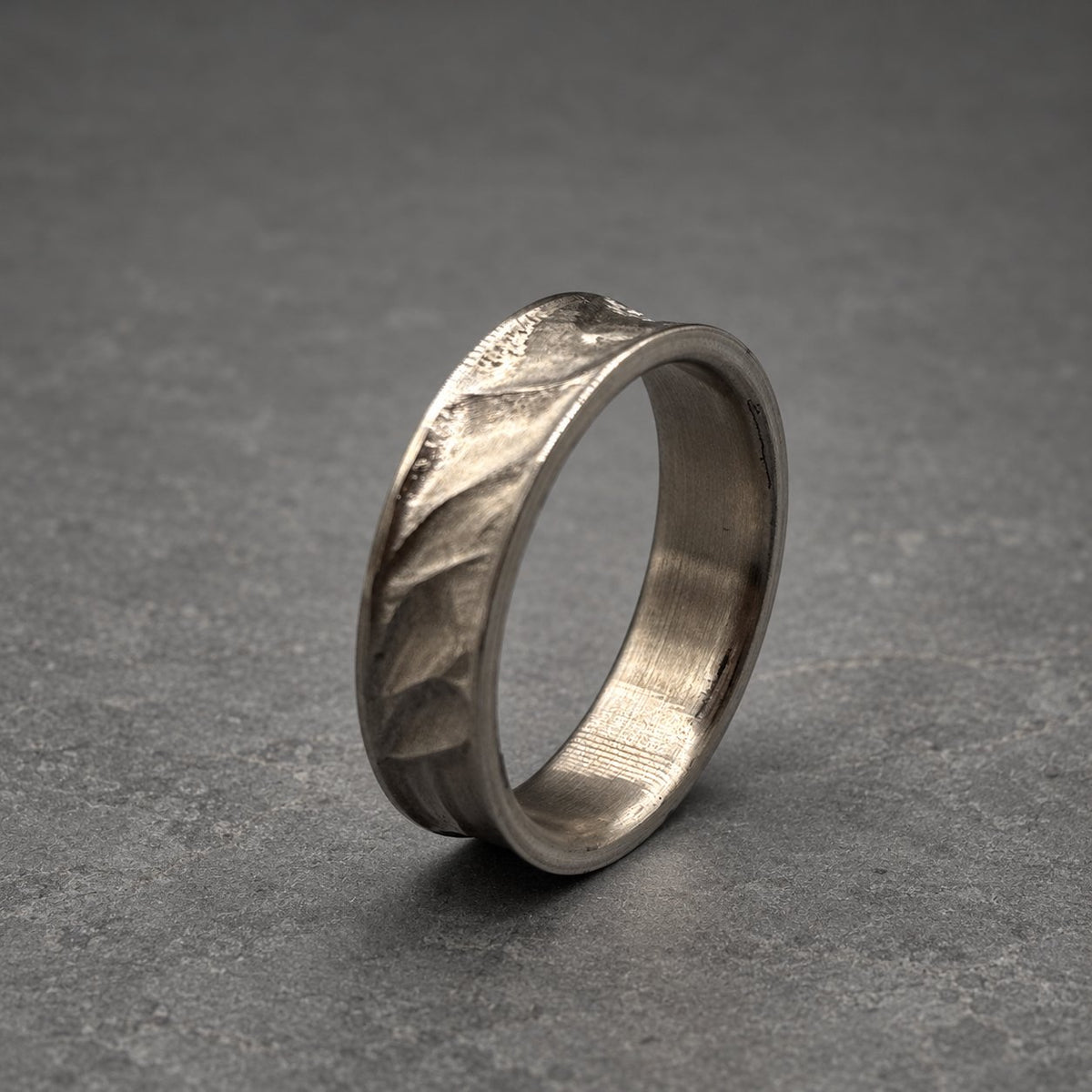 A silver ring with a textured, leaf-like pattern on the band. The ring has a slightly curved design and a smooth interior. The ring is shown against a gray background.
