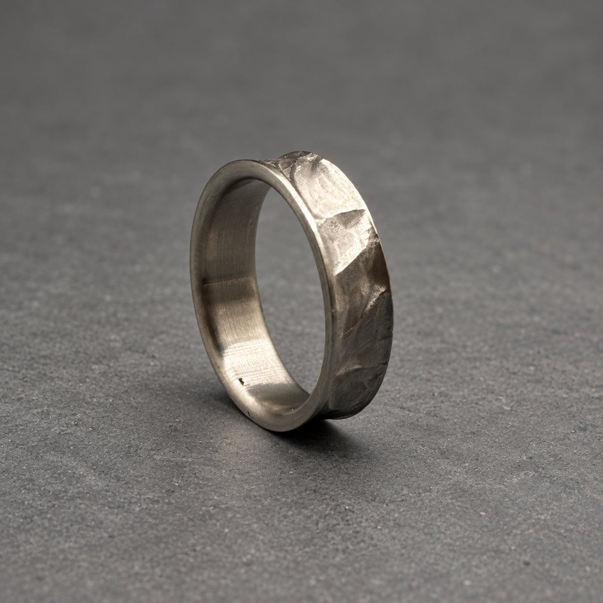 A silver ring with a textured, faceted surface. The ring has a band with a slightly curved design. The ring is set against a gray background.