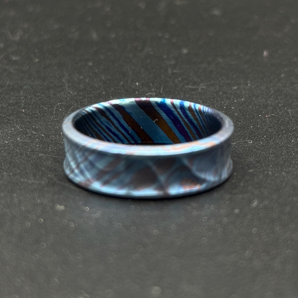 A blue ring with a patterned design. The ring has a textured surface with a repeating diamond pattern on the outside and a striped pattern on the inside. The colors include shades of blue, brown, and purple.