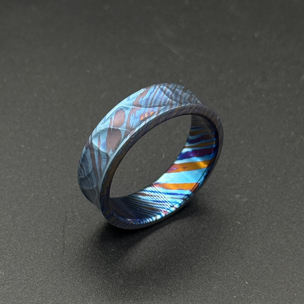 A patterned ring in shades of blue, purple, and orange. The ring has a textured surface and a band design. The ring is made of metal and is a striking piece of jewelry.
