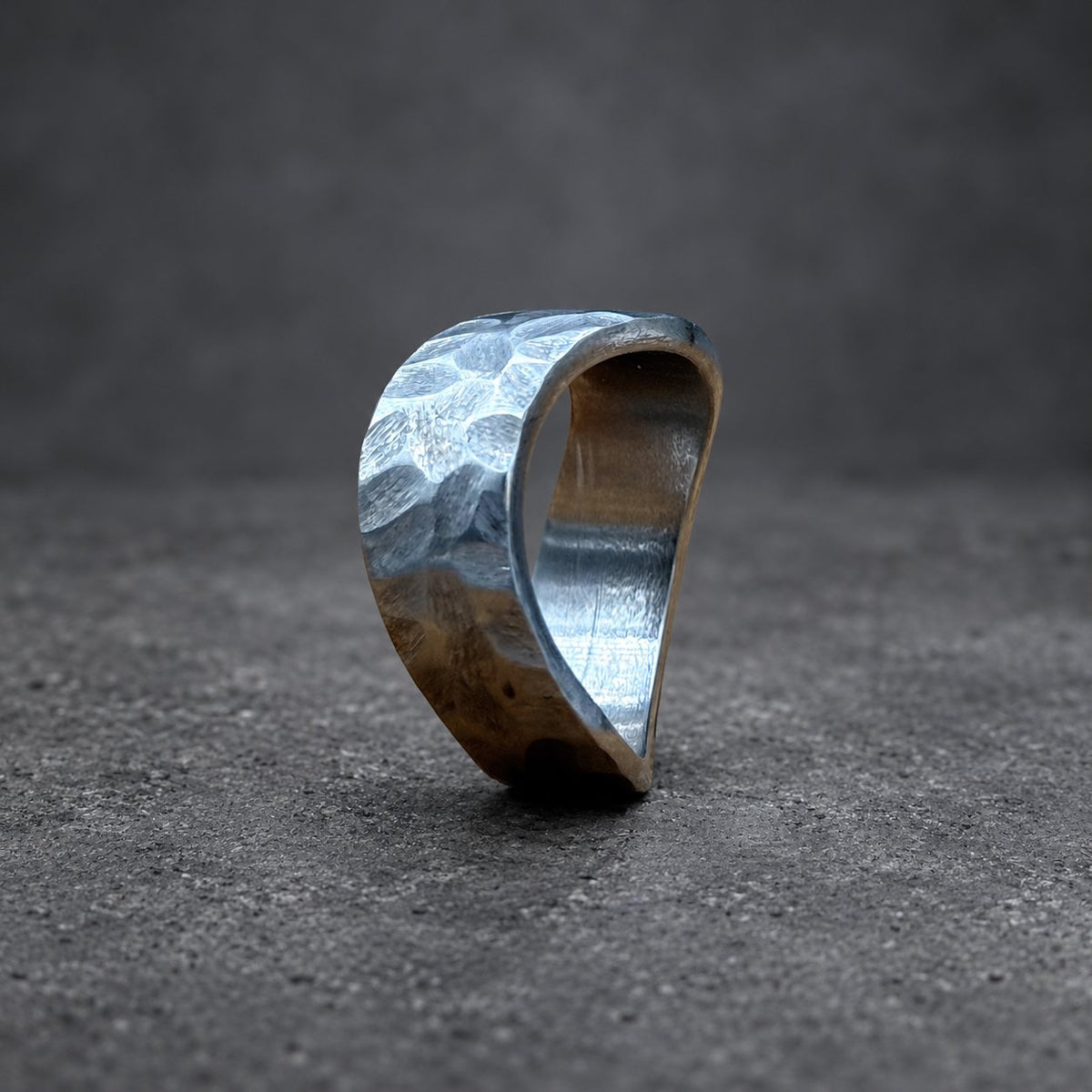 A hammered silver ring with a textured surface. The ring has a curved, open design and sits on a wooden surface. The ring's metal has a slightly reflective quality, and the background is blurred.