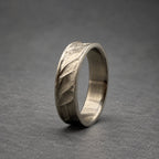 A silver ring with a textured leaf pattern. The ring has a brushed metal finish and a curved design. The ring is set against a gray background. The ring's design is reminiscent of nature.