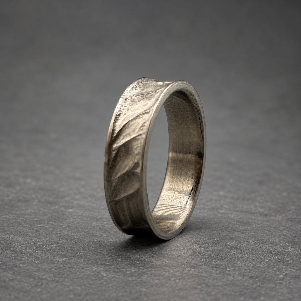 A silver ring with a textured leaf pattern. The ring has a brushed metal finish and a curved design. The ring is set against a gray background. The ring's design is reminiscent of nature.