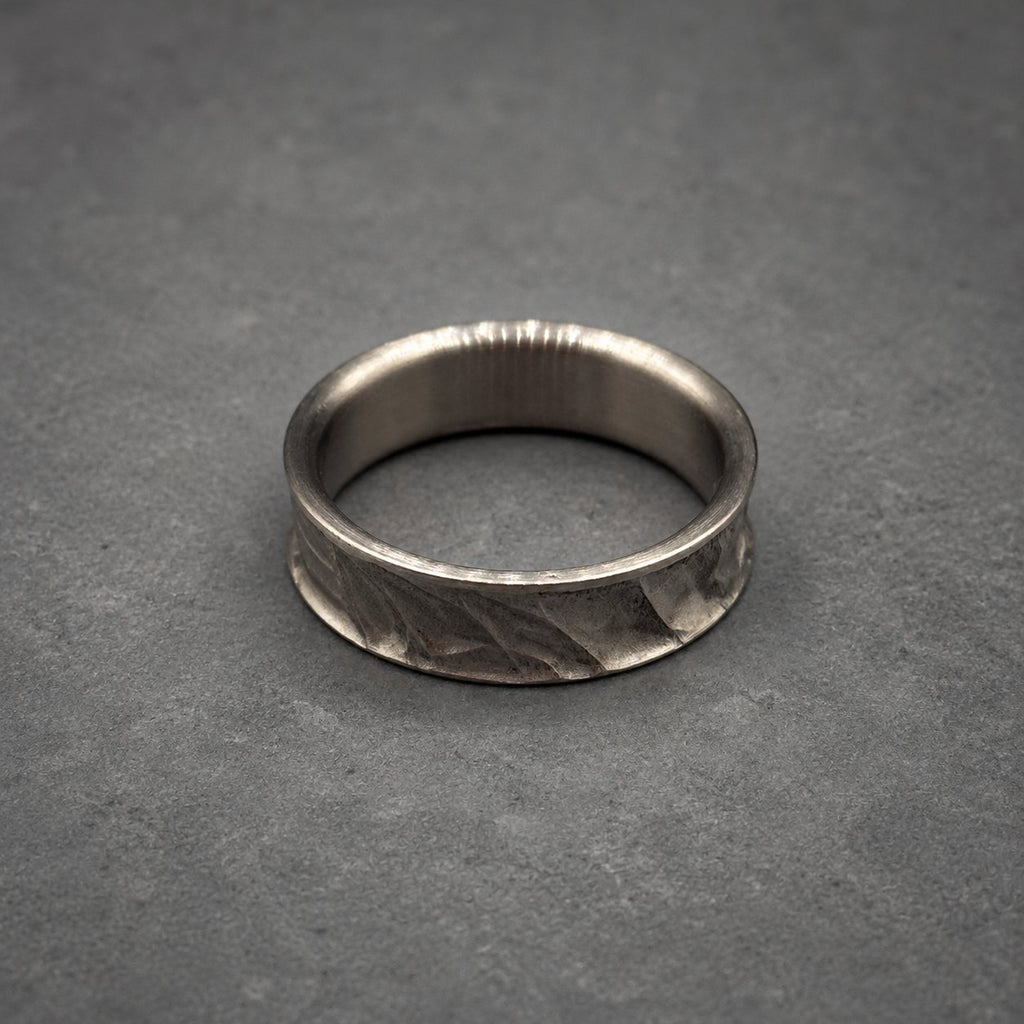 A silver ring with a textured band. The ring has a smooth inner surface and a patterned outer surface. The ring is displayed on a gray surface. Jewelry.