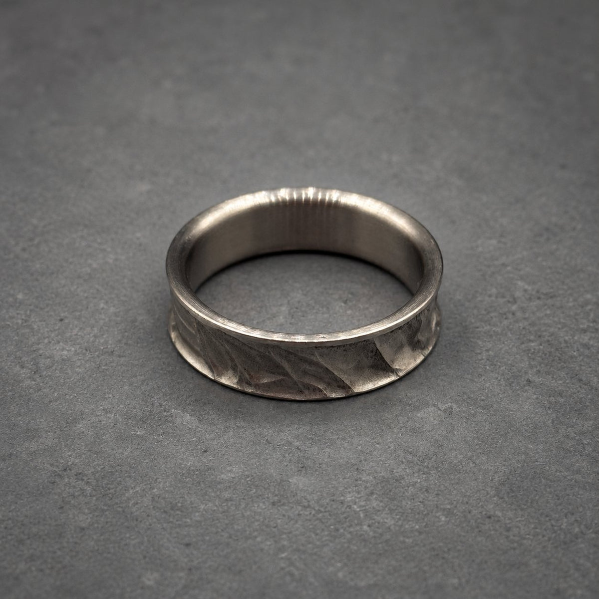 A silver ring with a textured band. The ring has a smooth inner surface and a patterned outer surface. The ring is displayed on a gray surface. Jewelry.