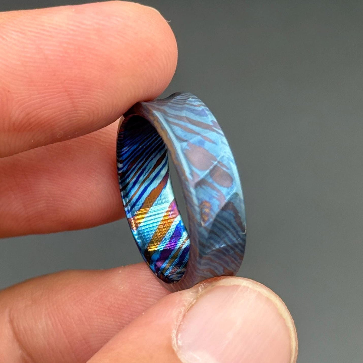A patterned ring in shades of blue and purple. The ring's design features a mix of colors and textures. The ring is held between two fingers. The ring's design is unique.