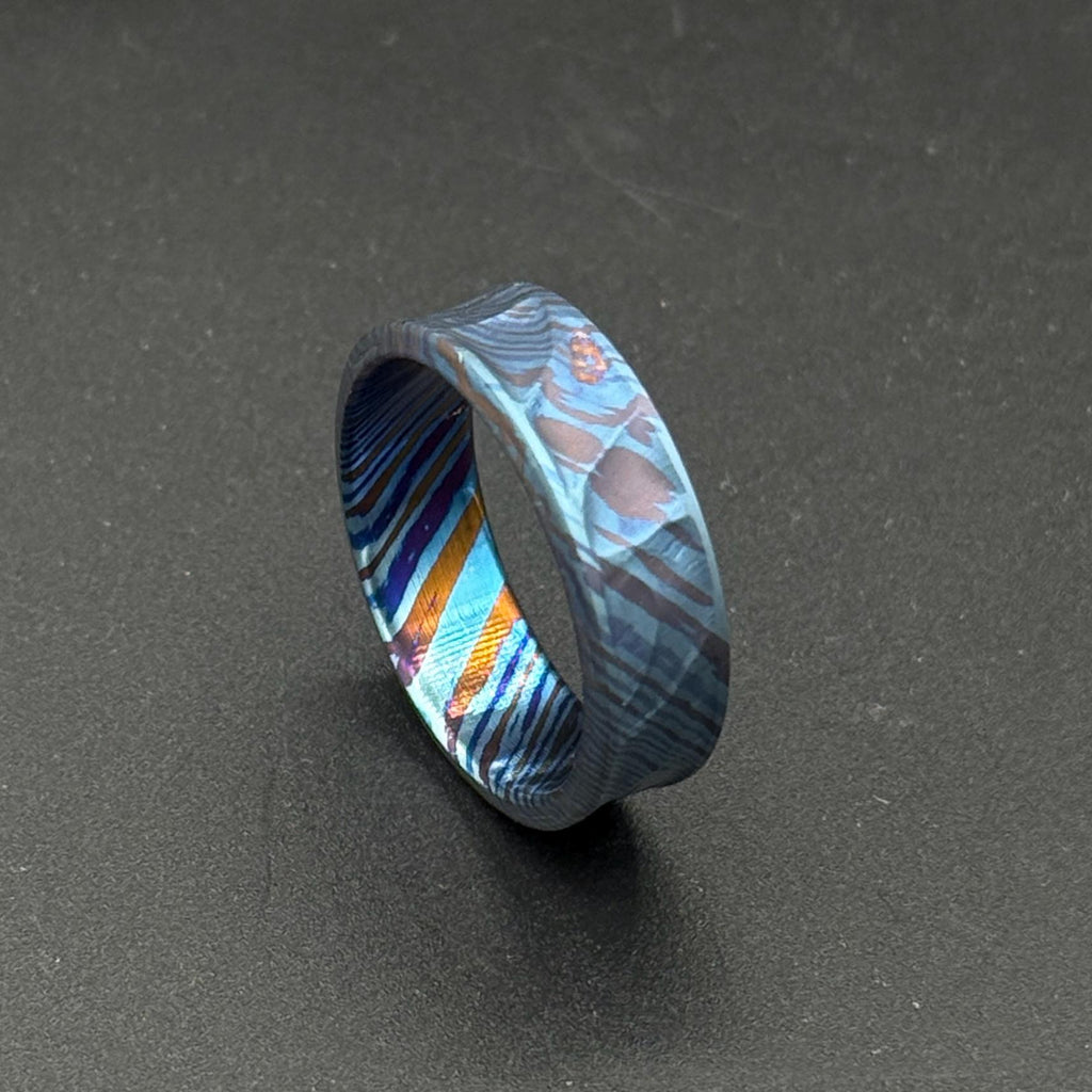 A patterned ring in shades of blue, purple, and orange. The ring's design is intricate and eye-catching, with a modern aesthetic. The ring is suitable for everyday wear or special occasions, with a unique and stylish appearance.