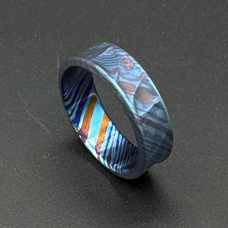 A patterned ring in shades of blue, orange, and purple. The ring has a textured surface with a mix of straight and curved lines. The ring is a piece of jewelry. The ring's design is eye-catching and unique.