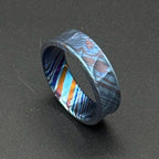 A patterned ring in shades of blue, orange, and purple. The ring has a textured surface with a mix of straight and curved lines. The ring is a piece of jewelry. The ring's design is eye-catching and unique.
