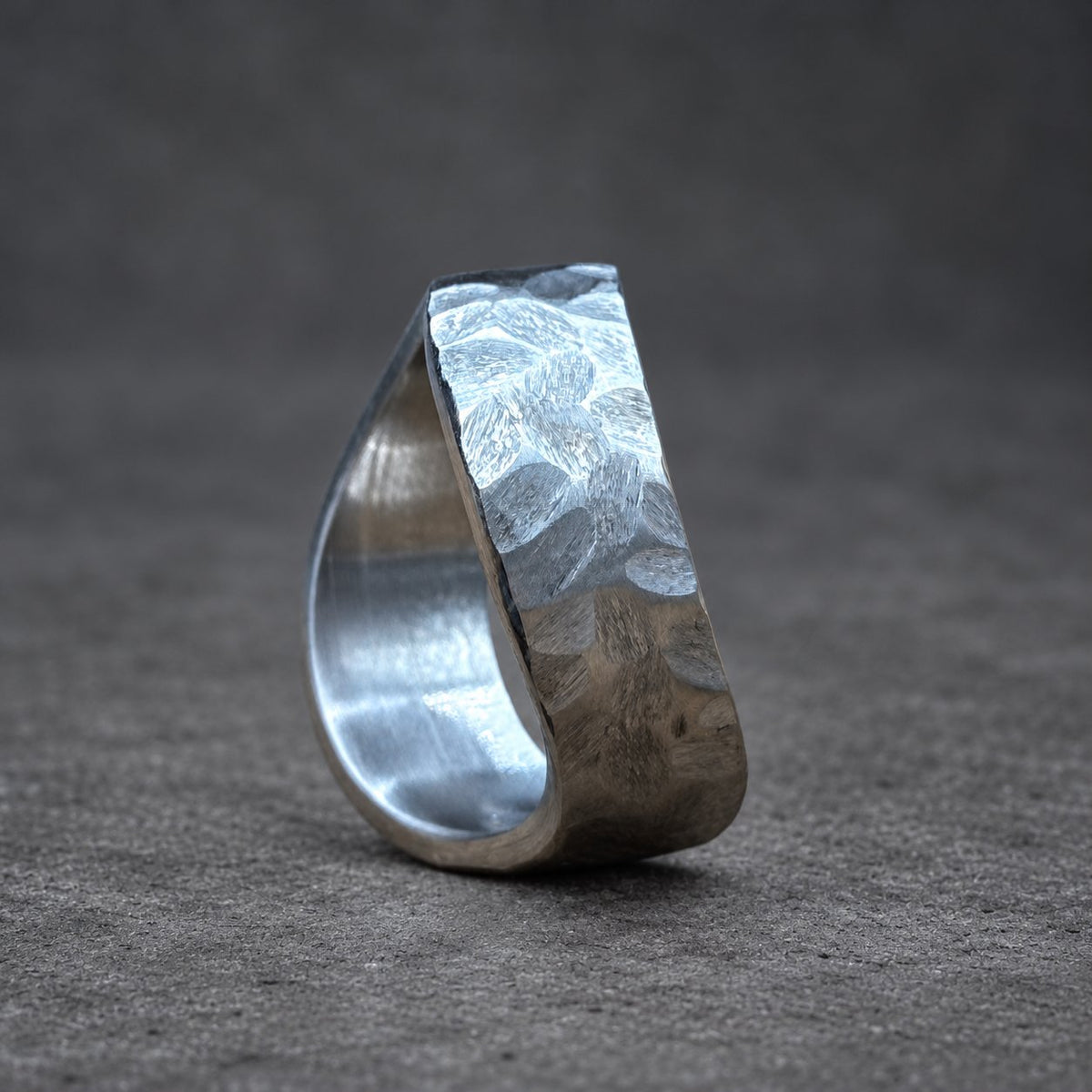 A silver ring with a hammered texture on the top section. The ring is teardrop-shaped, with a smooth, polished interior. The background is a blurred blue and wood grain.