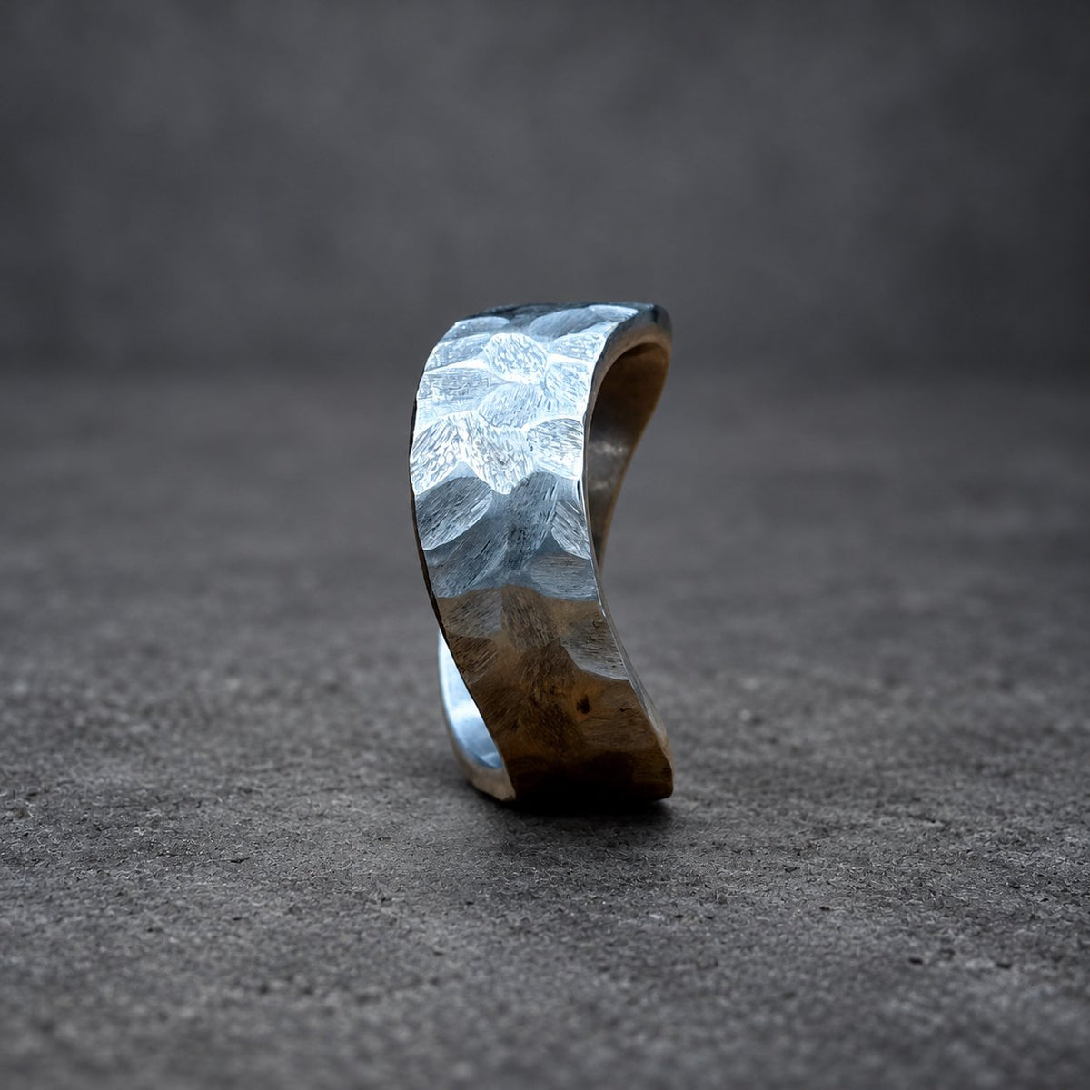 A silver ring with a hammered texture. The ring has a curved shape and is displayed on a wooden surface. The background is a blurred blue and brown.