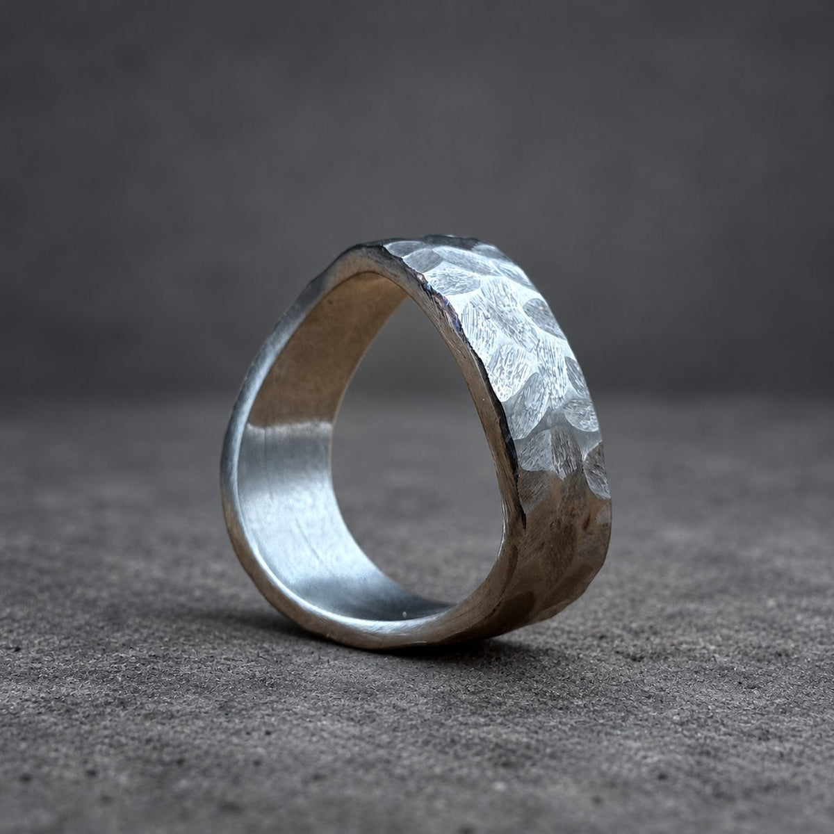 A silver ring with a hammered texture. The ring has a rounded band and a textured surface, creating a unique and rustic appearance. The ring is displayed on a wooden surface.