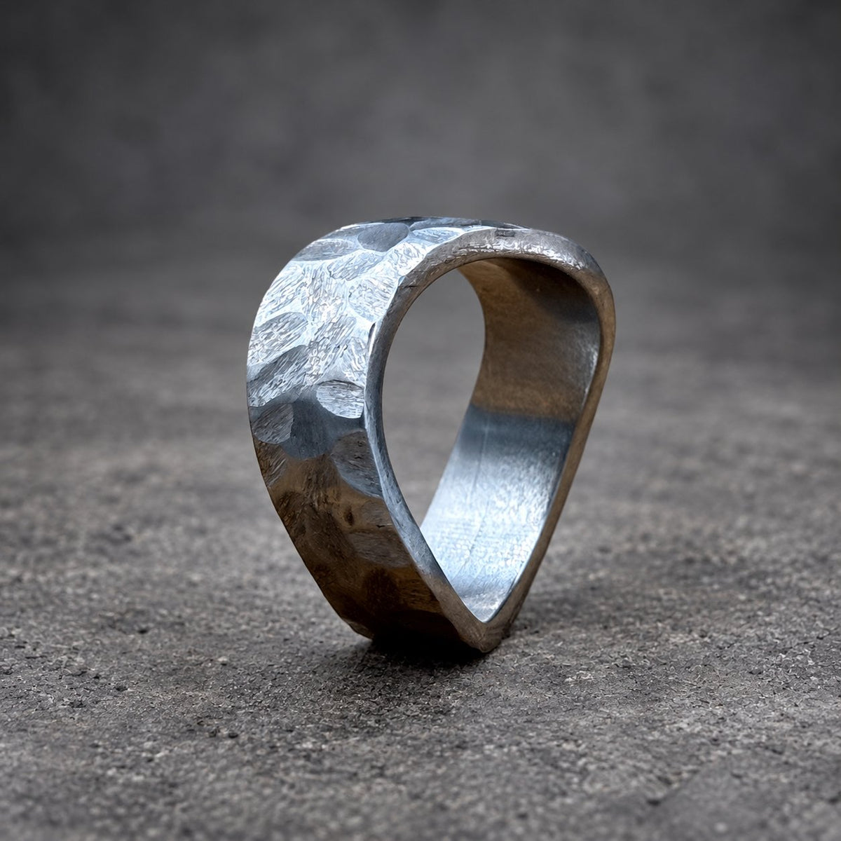 A silver-colored ring with a hammered texture. The ring has an unusual, open design. The ring is set against a wooden background. The ring's design is unique.