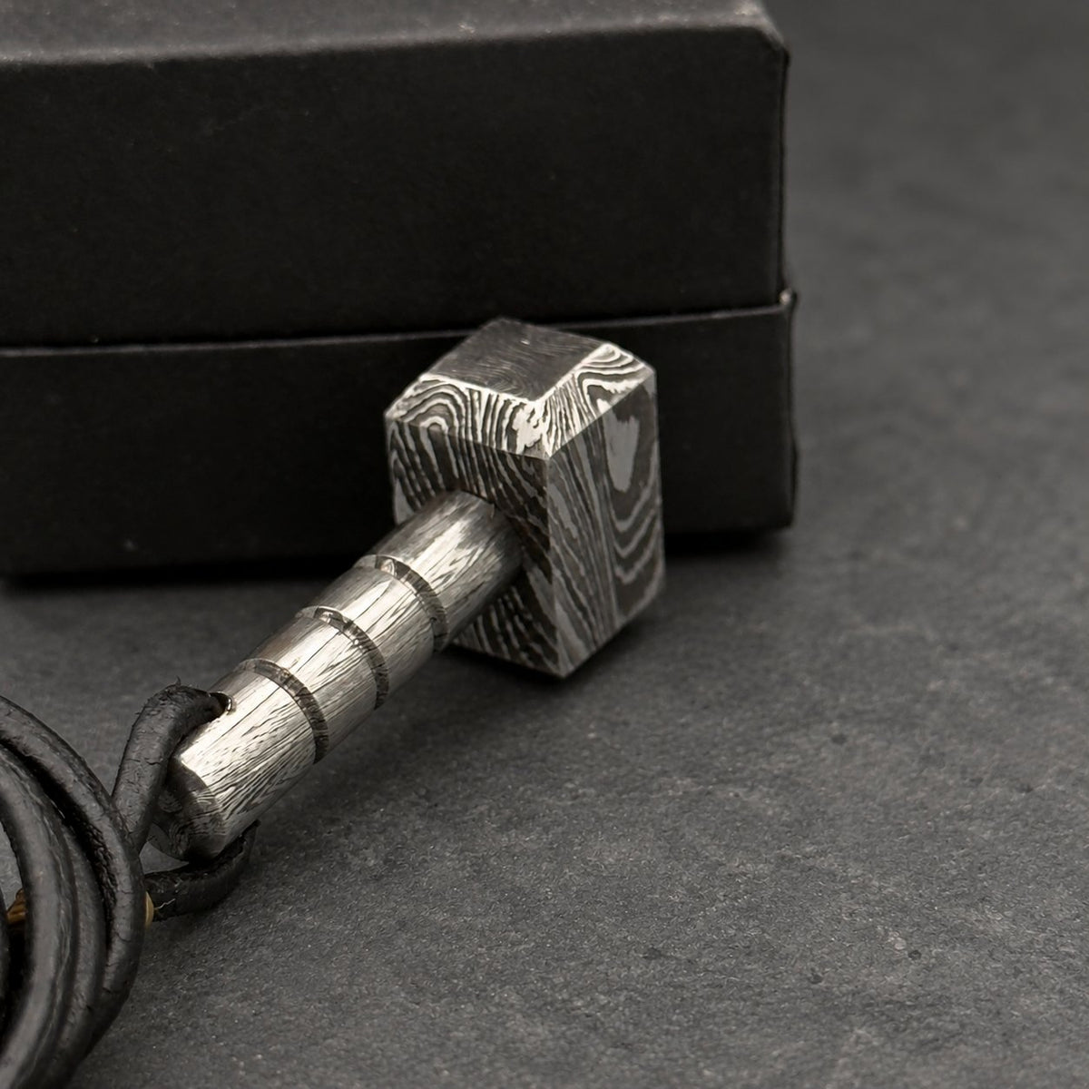Damascus steel Thor hammer keychain in front of a black gift box, cord coiled