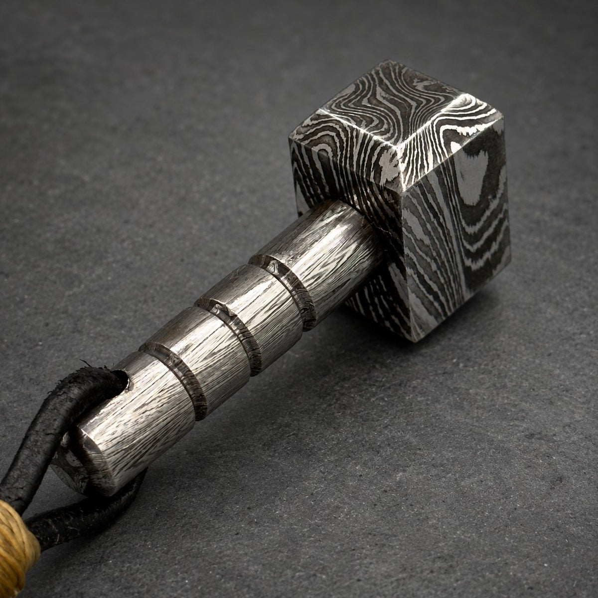 Damascus steel Thor hammer keychain on a gray surface, leather cord to one side
