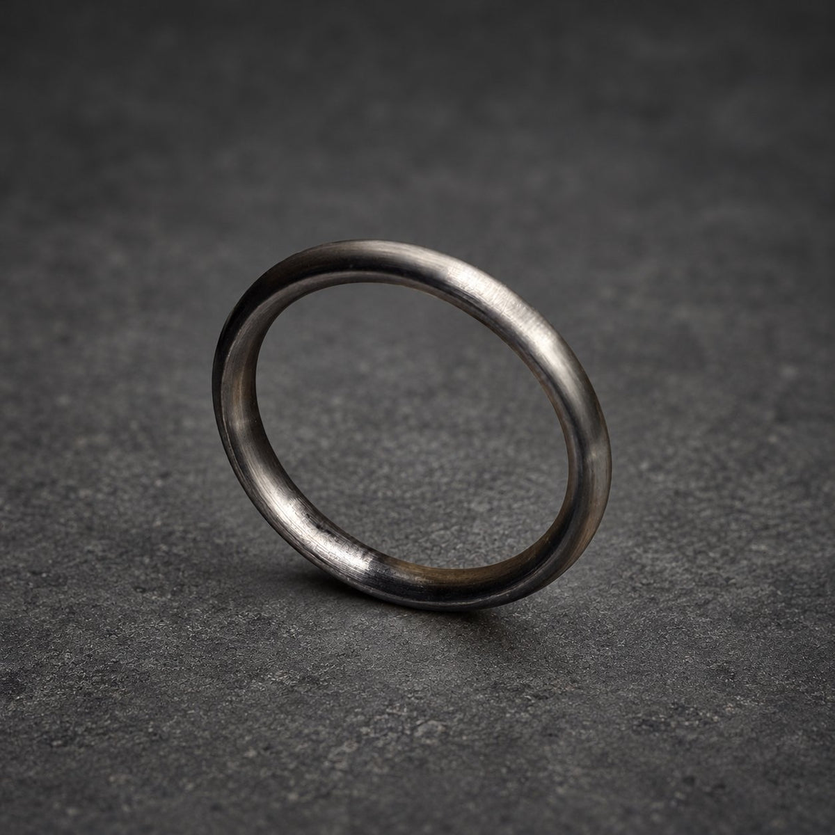 A close-up of a dark gray ring. The ring is made of metal and has a smooth, rounded surface. The ring is resting on a wooden surface. The ring is a simple, elegant design.