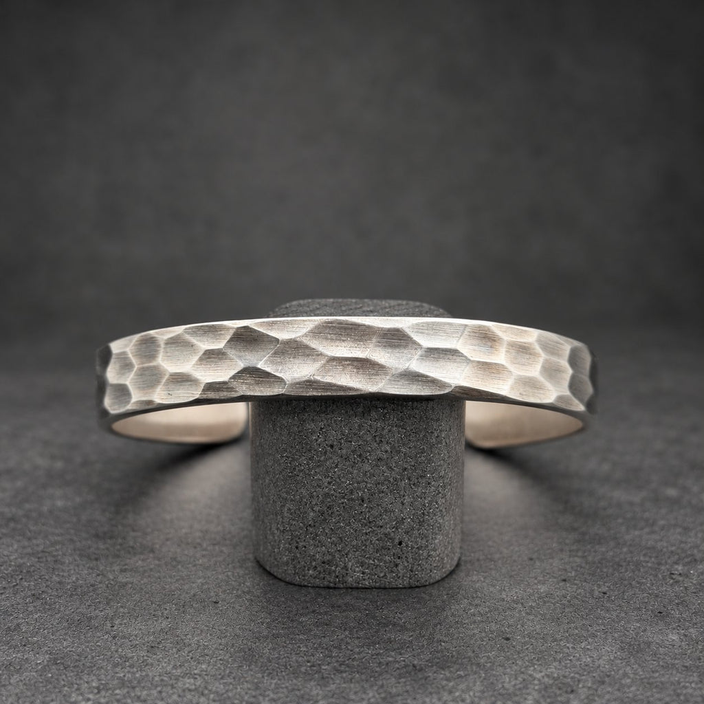 A silver cuff bracelet with a textured, hammered surface. The bracelet is displayed on a small wooden stand against a dark background. The cuff is open and has a geometric pattern.