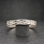 A silver cuff bracelet with a textured, hammered surface. The bracelet is displayed on a small wooden stand against a dark background. The cuff is open and has a geometric pattern.