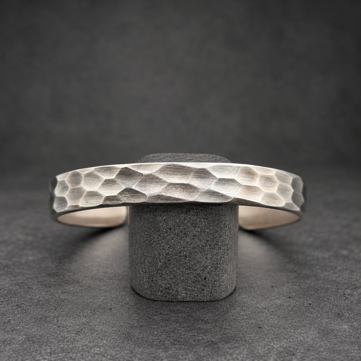 A silver cuff bracelet with a textured, hammered surface. The bracelet is displayed on a small wooden stand against a dark background. The cuff is open and has a geometric pattern.