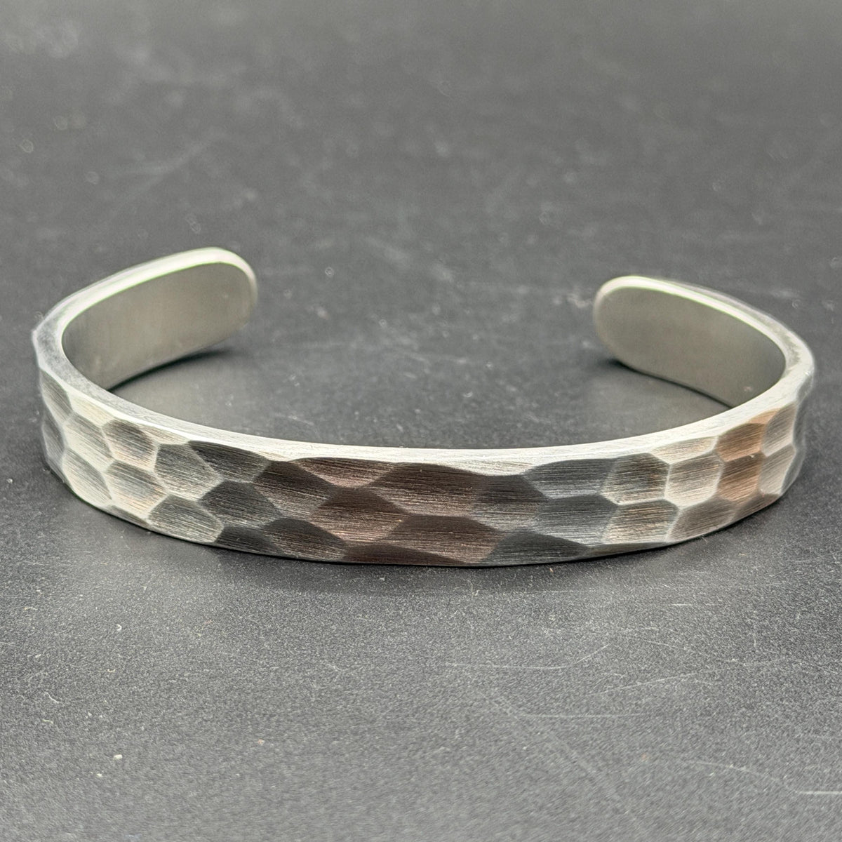 A silver cuff bracelet with a textured, hammered design. The open-ended bracelet has a rounded shape and geometric indentations. The bracelet is displayed on a dark surface.