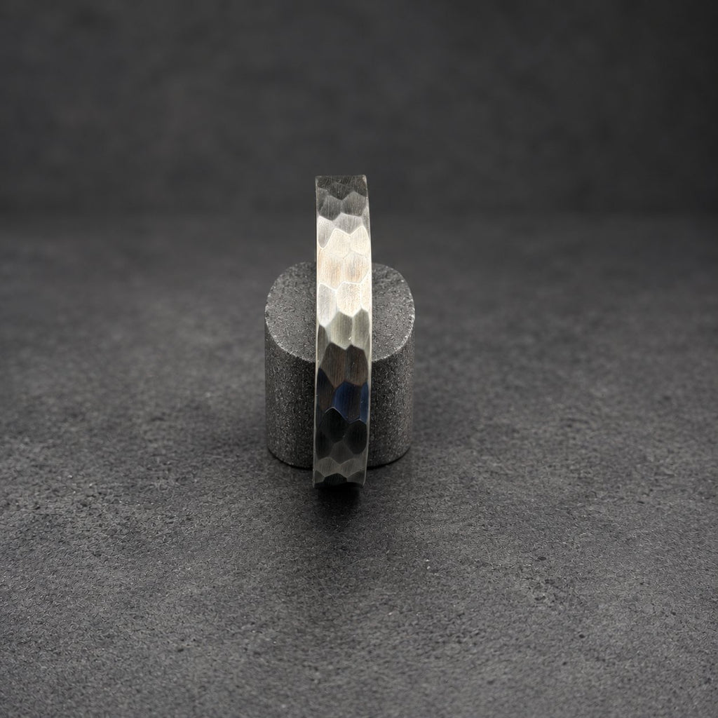 A silver metal bracelet with a textured, hexagonal pattern. The bracelet is displayed vertically on a small, cylindrical wooden stand against a dark background. The metal has a gradient of tones, from light silver to dark gray.
