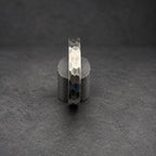 A silver metal bracelet with a textured, hexagonal pattern. The bracelet is displayed vertically on a small, cylindrical wooden stand against a dark background. The metal has a gradient of tones, from light silver to dark gray.