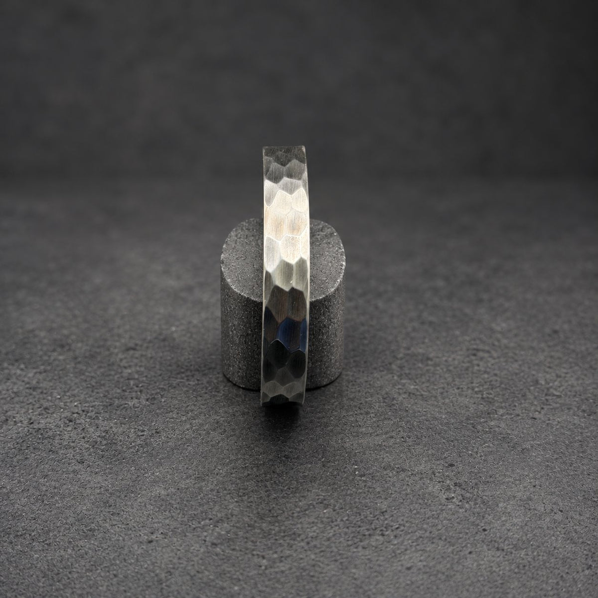 A silver metal bracelet with a textured, hexagonal pattern. The bracelet is displayed vertically on a small, cylindrical wooden stand against a dark background. The metal has a gradient of tones, from light silver to dark gray.
