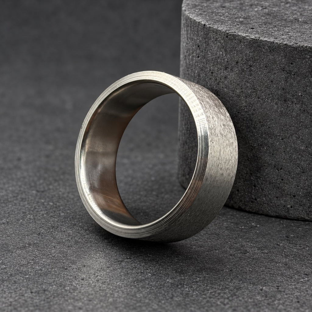 A wide, brushed silver ring. The ring has a textured surface and a smooth interior. It is displayed on a dark surface next to a piece of wood.