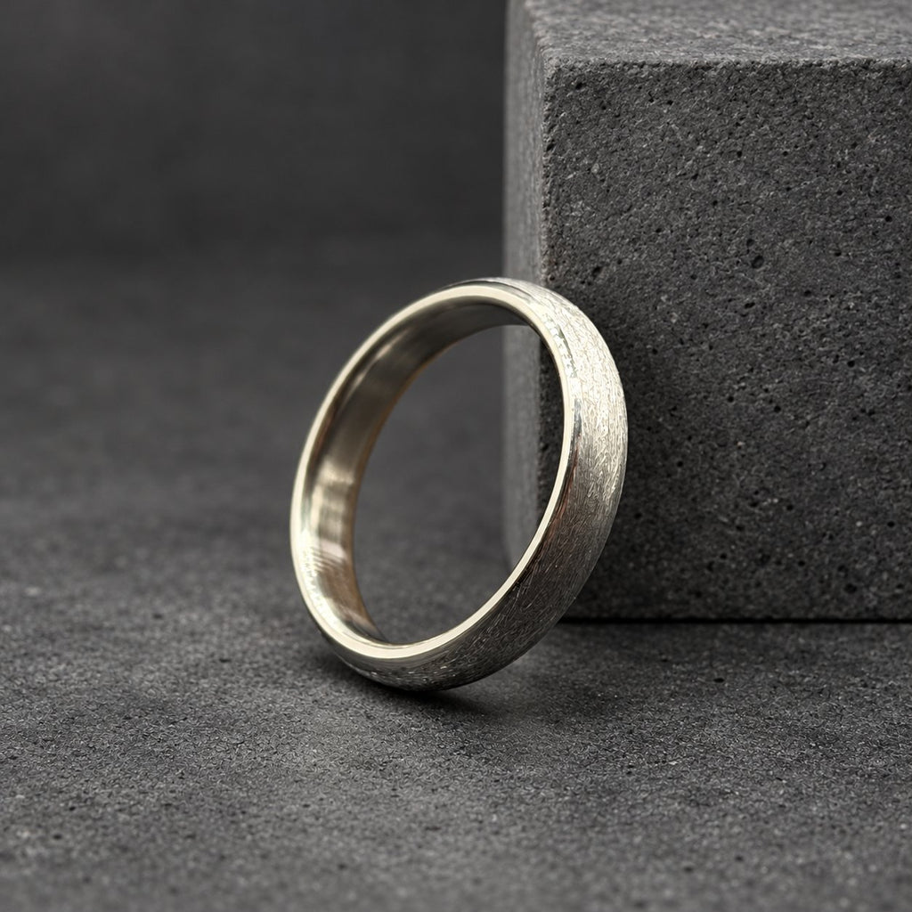 A silver ring with a textured, brushed finish. The ring is circular and sits on a dark surface, with a wooden block in the background. The ring's design is simple and elegant, suitable for everyday wear.