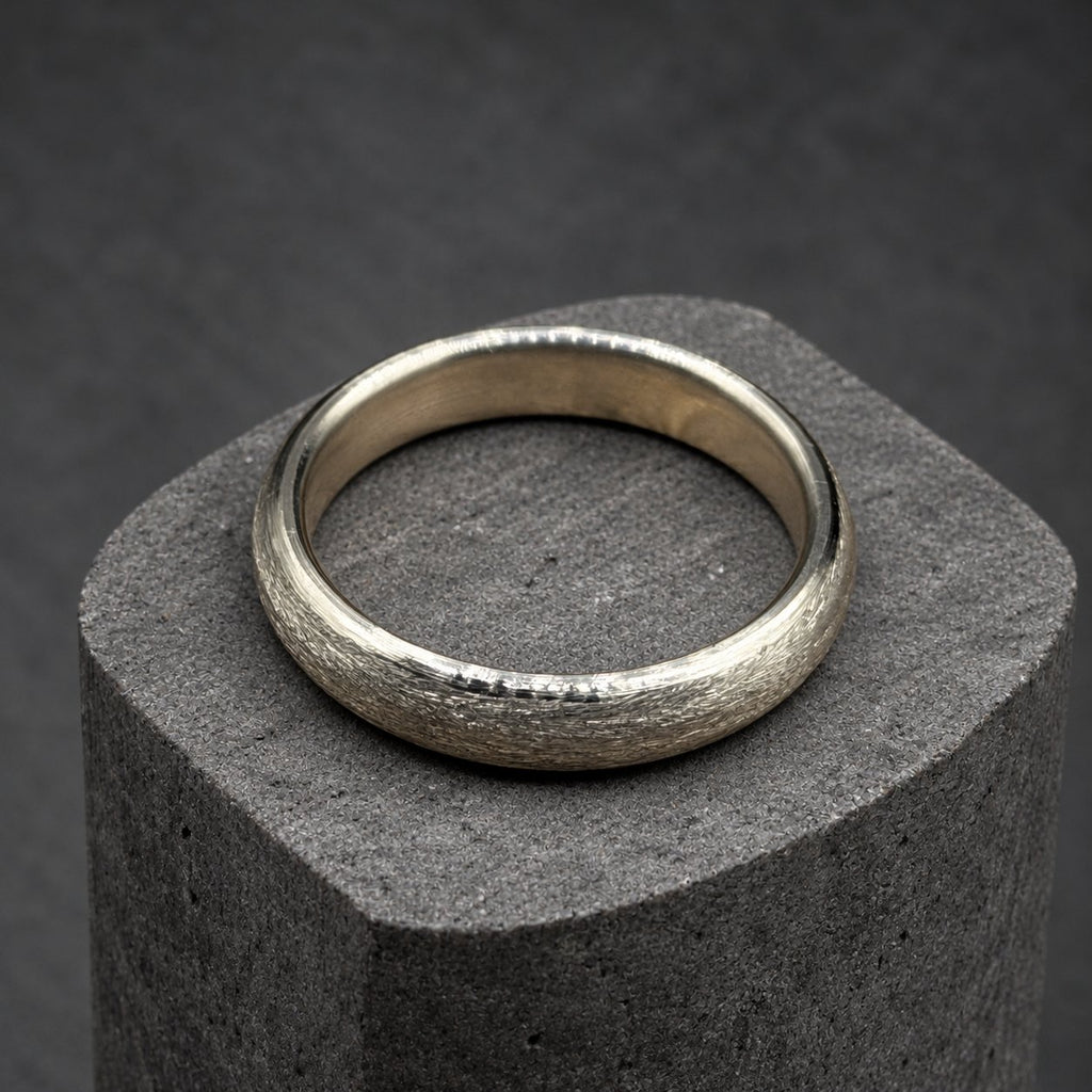 A silver ring with a textured surface sits on a wooden block. The ring has a simple band design and a matte finish. The wooden block has a natural wood grain pattern. The background is dark.
