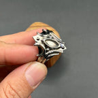 A silver ring with an elaborate, shield-shaped design. The ring has a central oval and multiple pointed edges, creating a bold and detailed aesthetic. The ring is held in a hand.