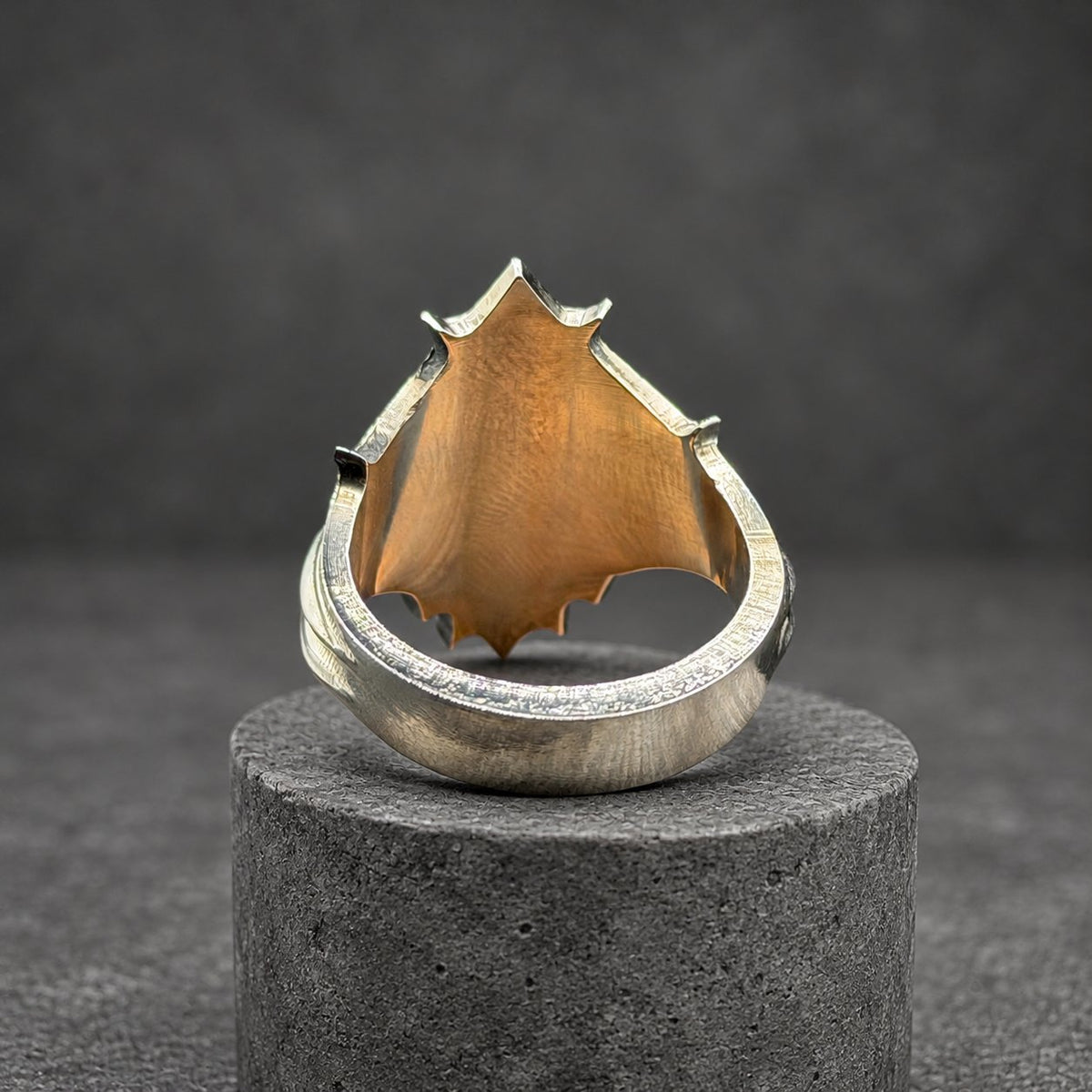A silver ring with a shield-shaped face. The ring's face has a dark, textured center and a pointed top. The band has a detailed design. The ring is displayed on a wooden block.