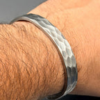 A silver cuff bracelet with a hammered texture. The bracelet features a hexagonal pattern and is worn on a wrist. The metal has a matte finish and reflects light.