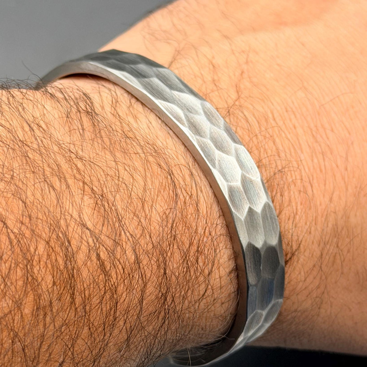 A silver cuff bracelet with a hammered texture. The bracelet features a hexagonal pattern and is worn on a wrist. The metal has a matte finish and reflects light.
