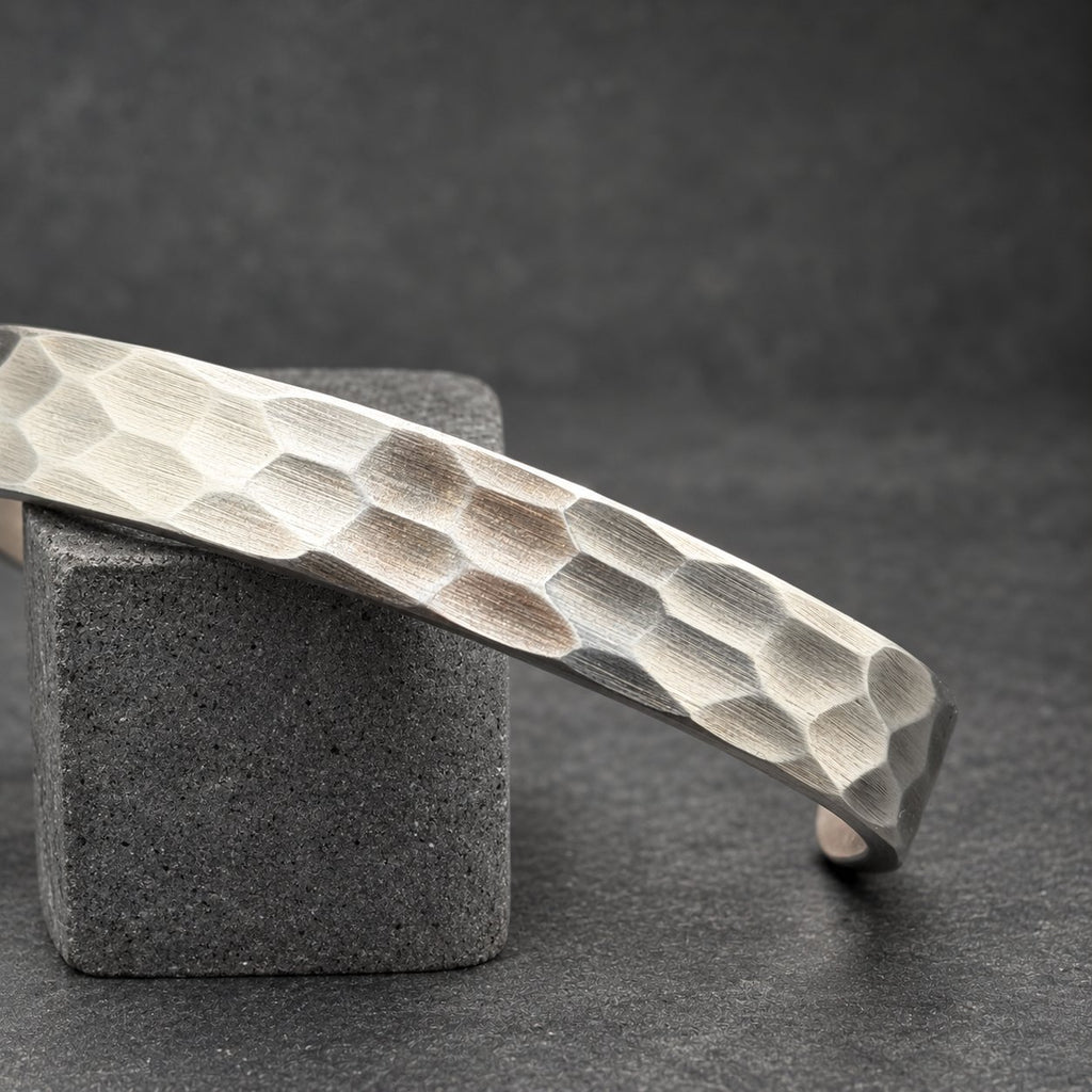 A silver cuff bracelet with a textured, honeycomb design. The bracelet is displayed on a small, wooden cylindrical object. The dark background accentuates the bracelet's metallic finish. A simple, modern piece of jewelry.