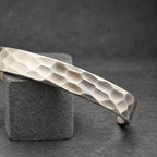 A silver cuff bracelet with a textured, honeycomb design. The bracelet is displayed on a small, wooden cylindrical object. The dark background accentuates the bracelet's metallic finish. A simple, modern piece of jewelry.