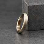 A silver ring with a textured finish. The ring is positioned in front of a wooden block with orange and brown hues. The ring has a plain band design.