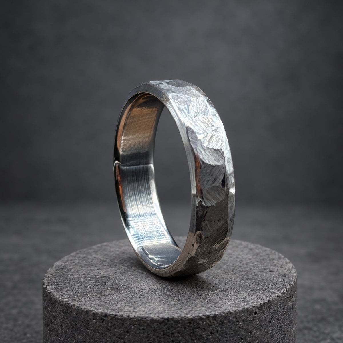 A silver ring with a hammered texture. The ring is presented on a wooden stand. The band has a slightly rounded shape. The ring's finish gives it a unique, handcrafted appearance.