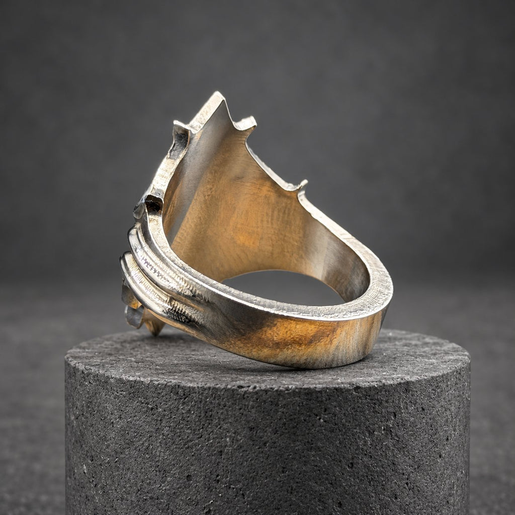 A silver ring with a distinctive, angular design. The ring has a textured surface and a prominent, pointed top, with curved lines on the side. The ring is displayed on a wooden block, highlighting its intricate details.