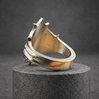 A silver ring with a distinctive, angular design. The ring has a textured surface and a prominent, pointed top, with curved lines on the side. The ring is displayed on a wooden block, highlighting its intricate details.